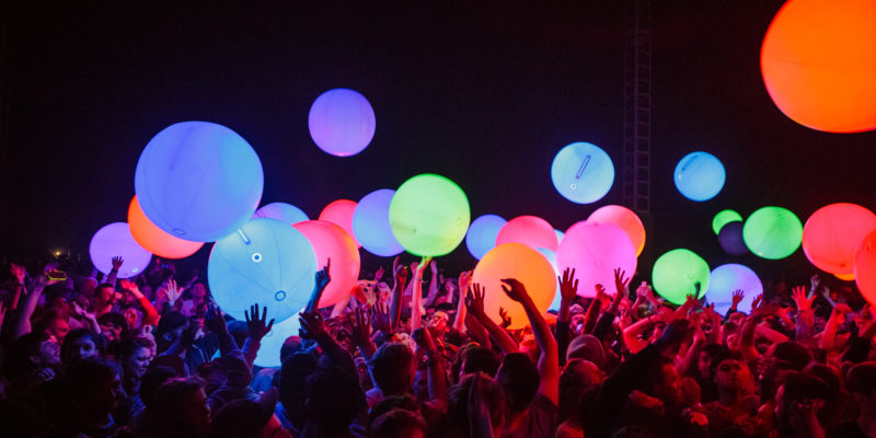 A large group of young people crowded together with their hands raised above their heads, with colourful illuminated spheres floating above them against a dark background