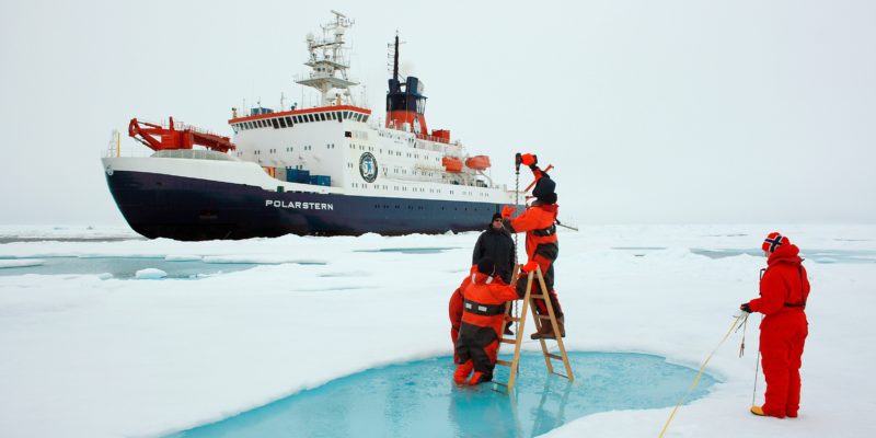 Four people setting up equipment, one standing on a ladder on a patch of ice, with a ship in the background