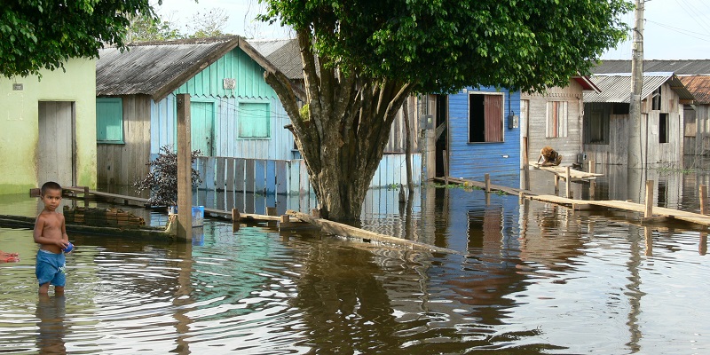 A flooded village, showing water rising up the outside walls of residential buildings and trees, with a child stood in knee-deep water