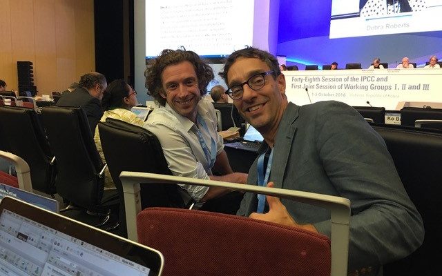 Professors Piers Forster and James Ford smiling into the camera in a conference auditorium