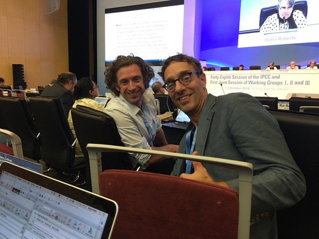 Professors Piers Forster and James Ford smiling into the camera in a conference auditorium