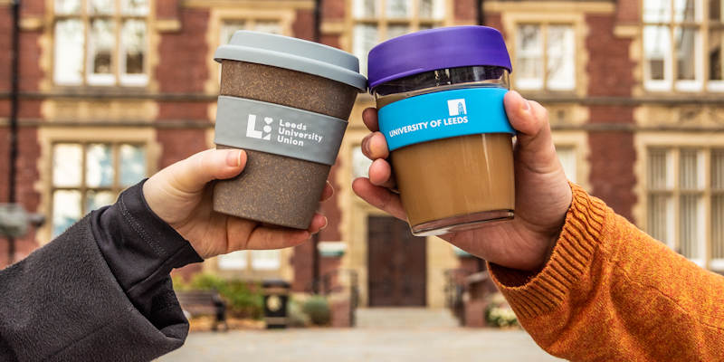 Close up of two hands holding reusable coffee cups with University of Leeds logos