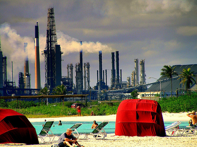 People relaxing at a poolside, with an oil refinery working in the back