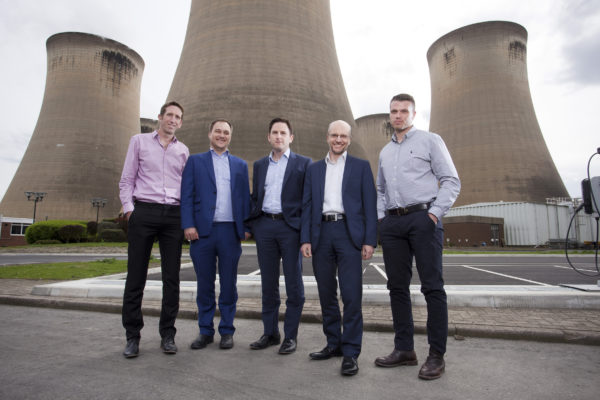 A group of five men wearing suits posing in front of some cooling towers