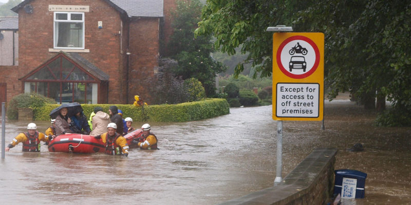 A goup of people up to their chests in water, dragging an inflatable dinghy through a flooded residential street, with a group of residents sitting in the boat