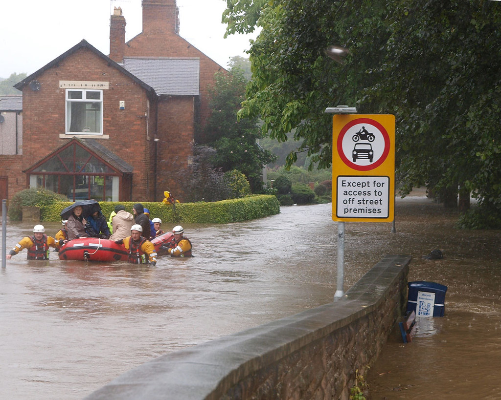A goup of people up to their chests in water, dragging an inflatable dinghy through a flooded residential street, with a group of residents sitting in the boat