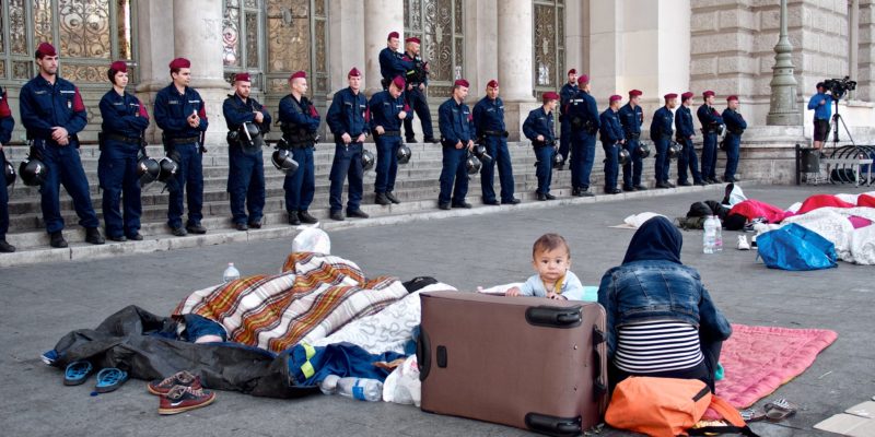 Families sitting outside an official building with luggage and children, surrounded by police and being filmed