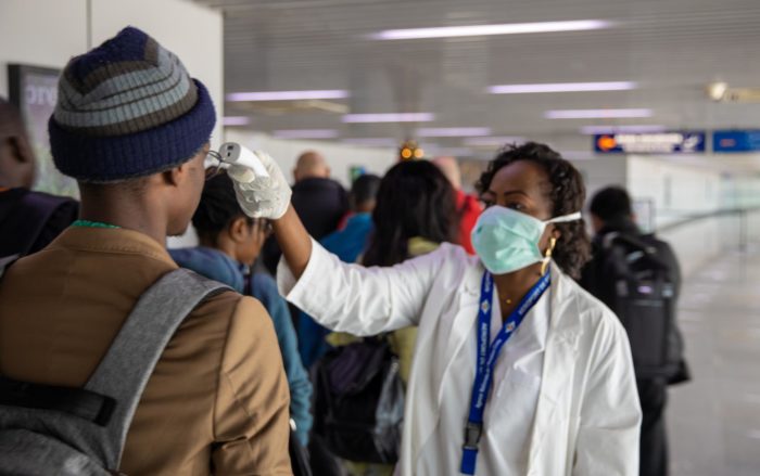 People standing in line at an airport having their temperature checked for COVID-19 by a medical practitioner with a facial mask on