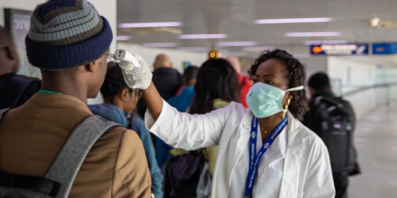 People standing in line at an airport having their temperature checked for COVID-19 by a medical practitioner with a facial mask on