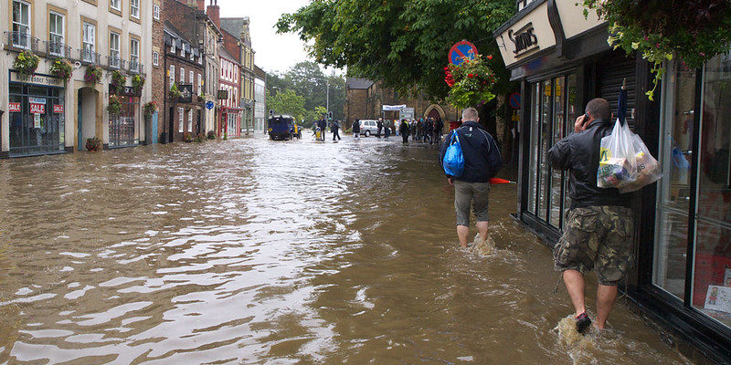 Shopping street in York, flooded to shin level on the people walking away and closest to the camera, and vehicles and more people in the distance