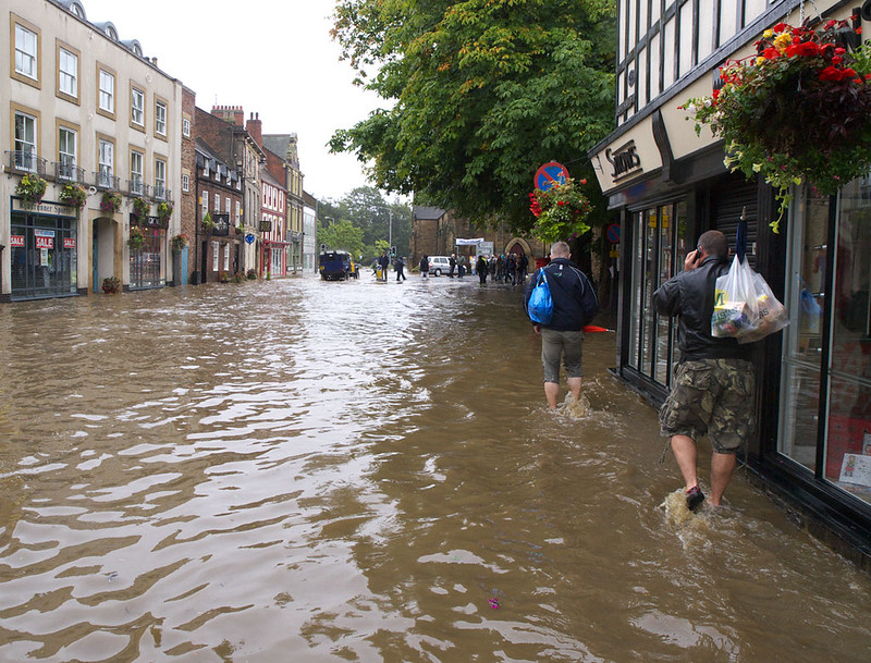 Shopping street in York, flooded to shin level on the people walking away and closest to the camera, and vehicles and more people in the distance