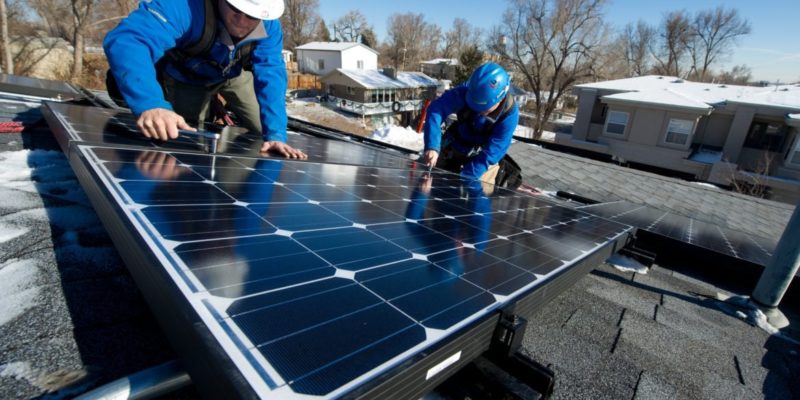 Two men in blue are installing a solar panel on the roof of a building on a sunny day