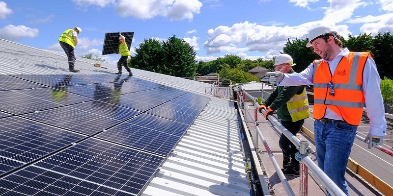 Men on a large roof direct others to install solar panels on a sunny day