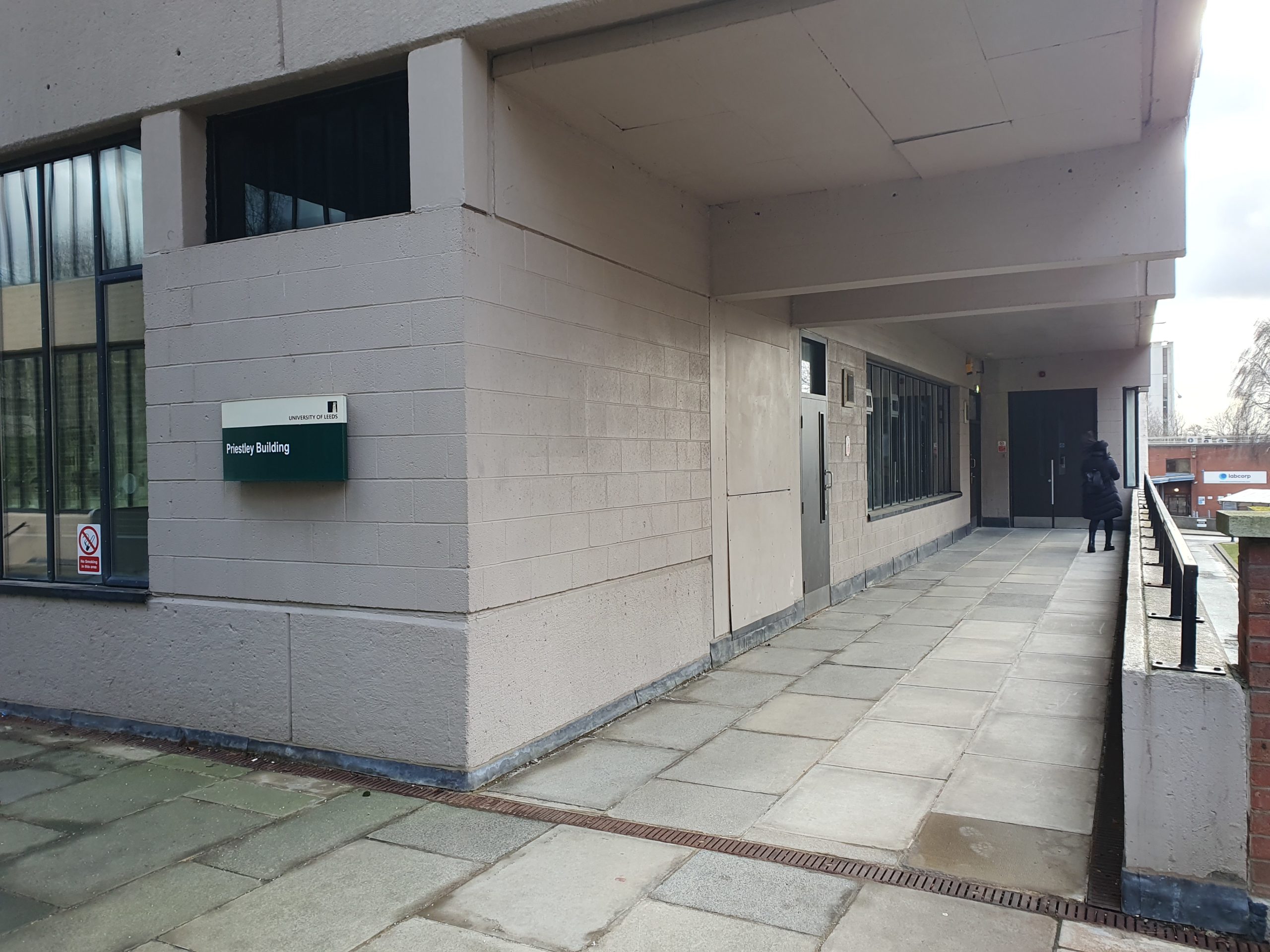 Entrance to the Priestley Building. There is a green sign reading "University of Leeds Priestley Building" to the left hand side of the entrance, with brown double doors leading into the building.