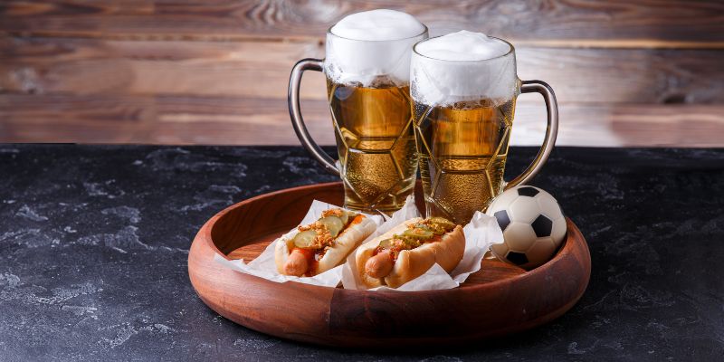 Two hot dogs on a wooden board with a mini-football and two beers.